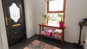 Een kamer met een zwarte deur, een raam met een kleurrijke gordijntje, een klein houten tafeltje met een bloevas en een stofzuiger tegen de muur.