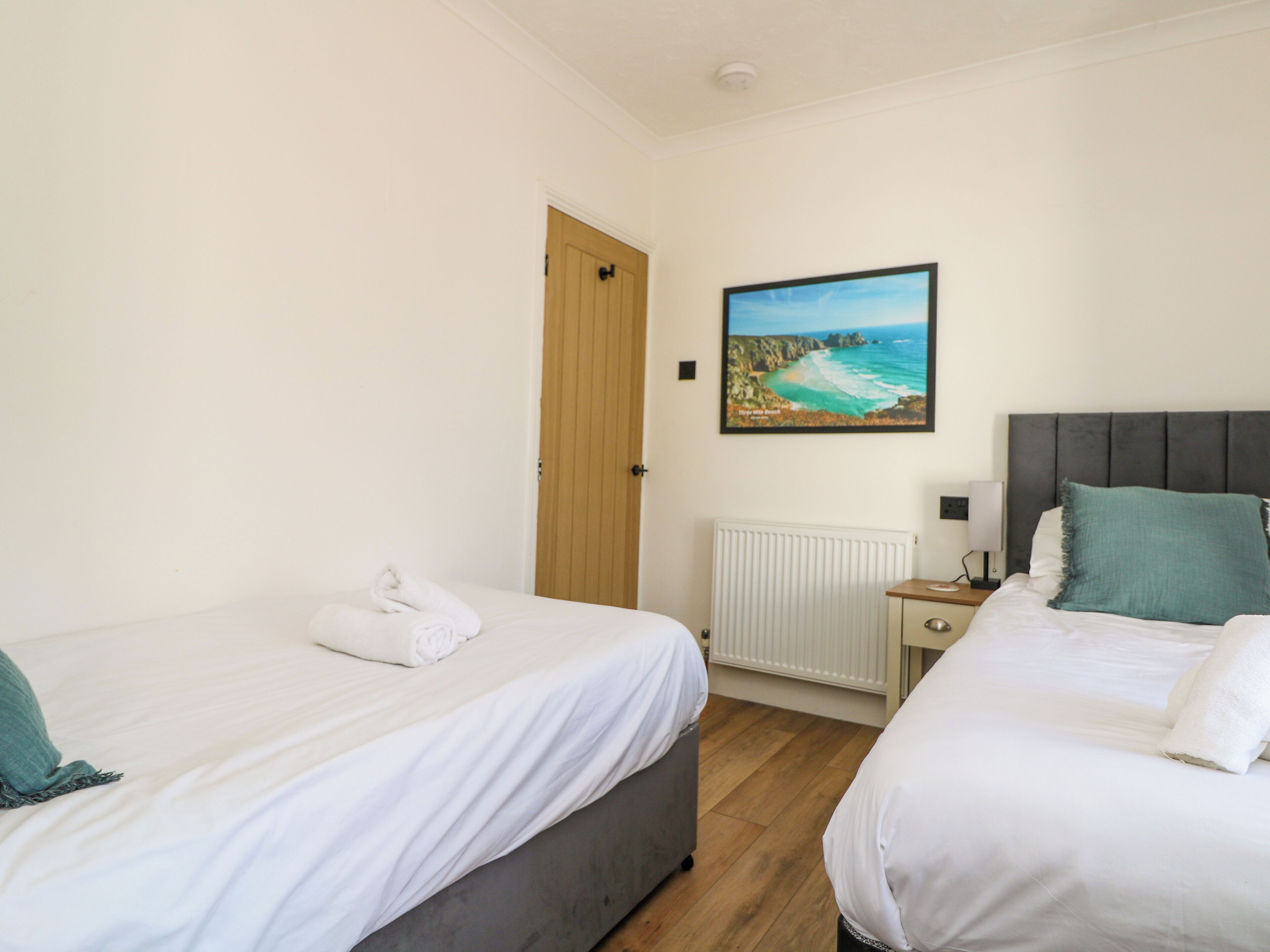 A hotel room with two beds, a wooden door, a painting of a coastal scene, a bedside table with a lamp, and a radiator.