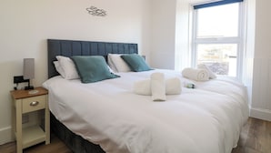 Una cama bien tendida con sábanas blancas y almohadillas color turquesa, una mesita de noche con una lámpara, y una ventana con vistas al exterior.