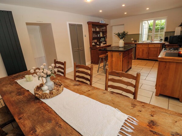 A dining area with a wooden table, chairs, and a white table runner. There is a kitchen area with wooden cabinets and a sink.