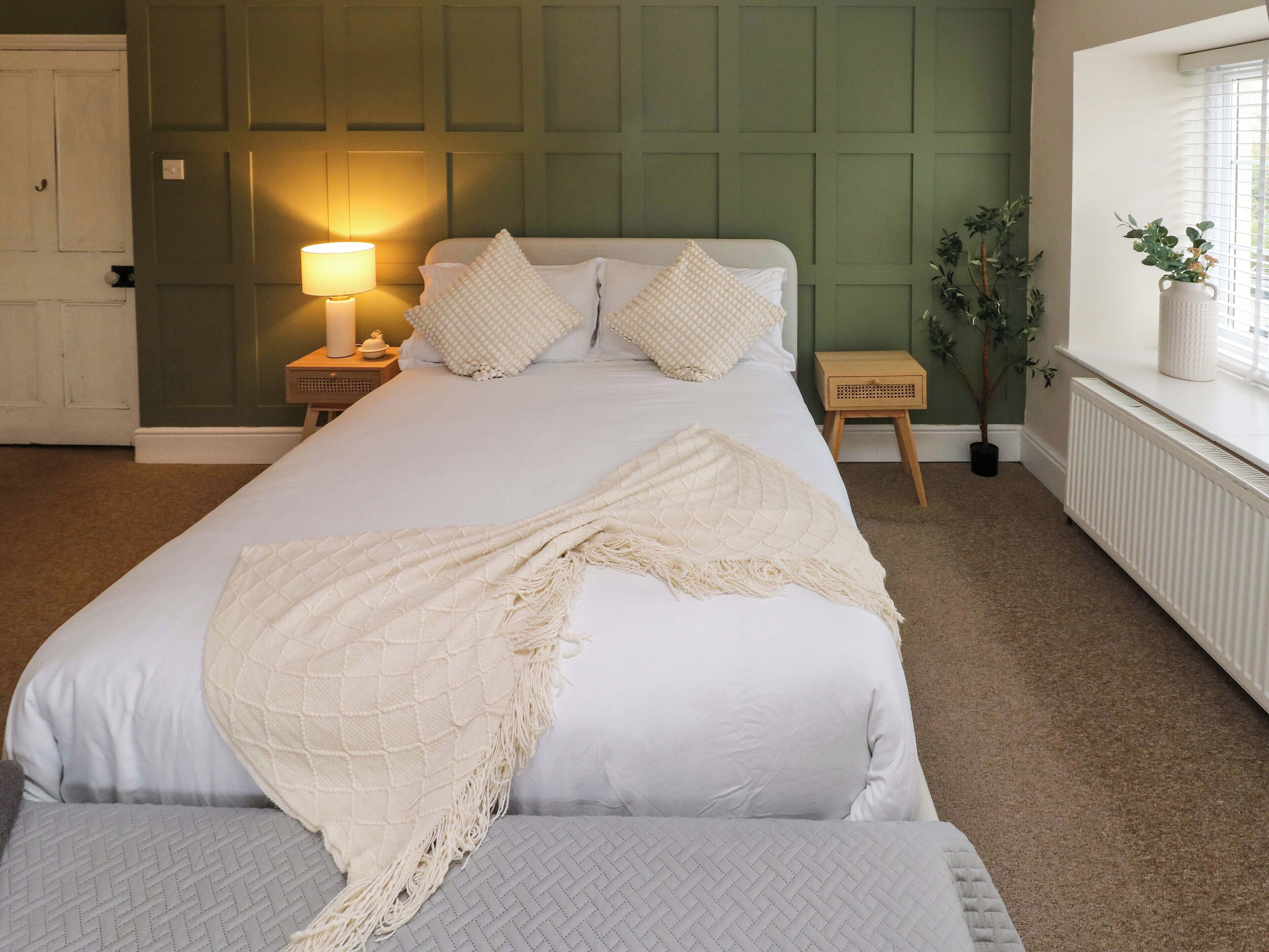 A neatly arranged bedroom with a bed, bedside tables, a lamp, and a window with a plant.