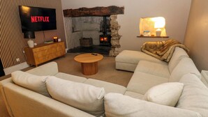 Een woonkamer met een modulaire bank, een houten salontafel, een stenen open haard, een televisie waarop 'NETFLIX' wordt weergegeven en een lamp op een plank.