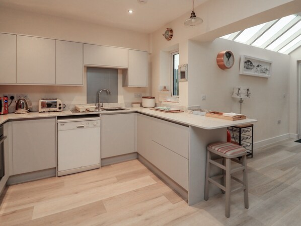 A modern kitchen with a dishwasher, a toaster, and a sink. There is a window above the sink and a clock on the wall.