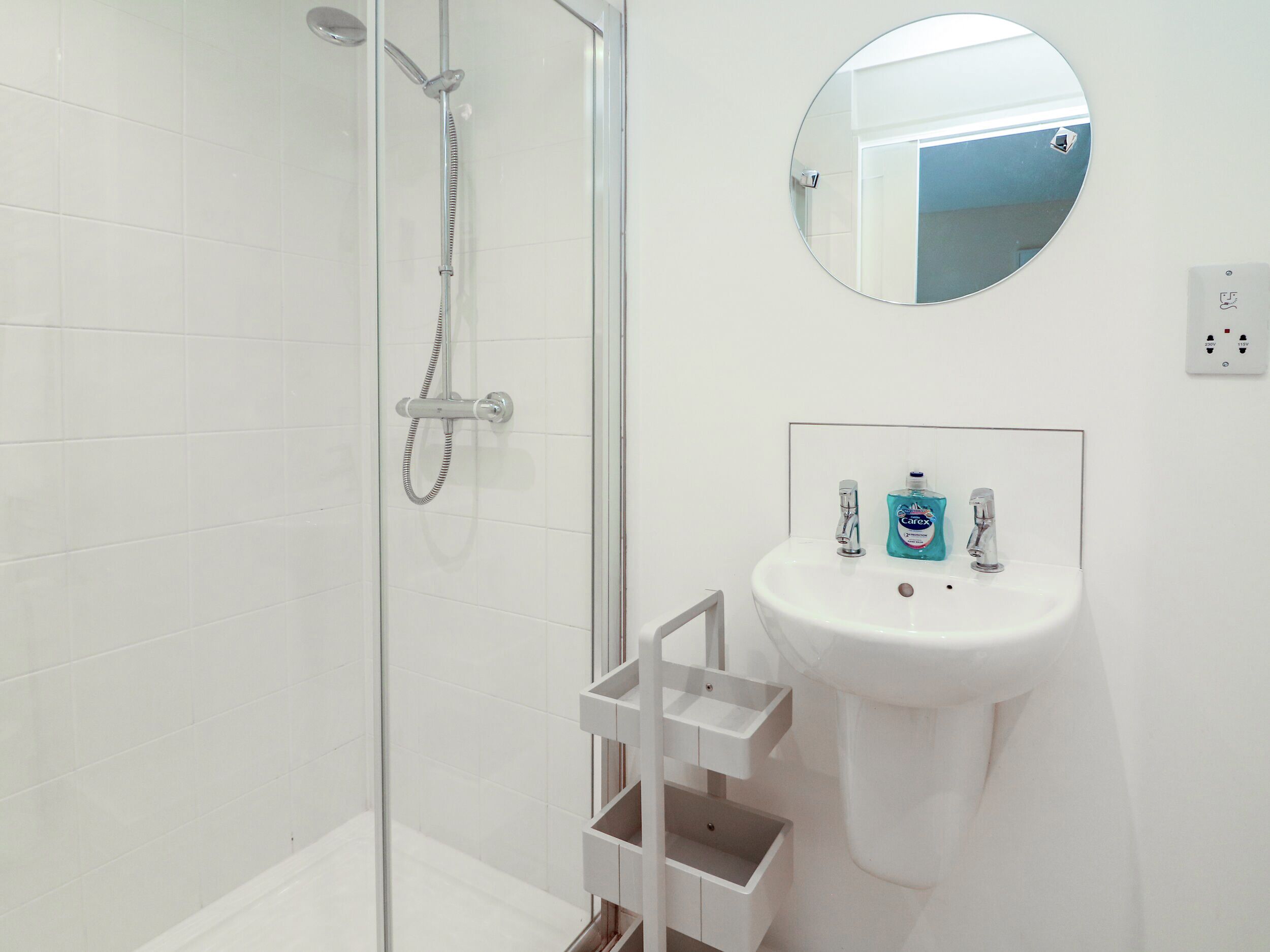 Een badkamer met een douche met glazen wand, een witte wastafel met een spiegel erboven en een fles Carex mondspoeling op de wastafel.