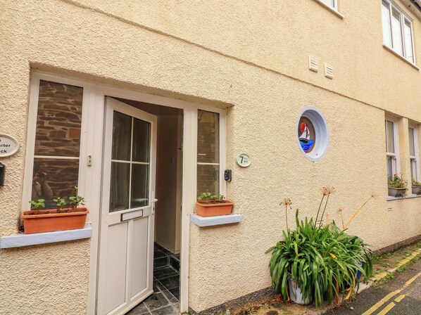 A building with a beige textured exterior, a door with a window, and a small circular window with a decorative element. There are potted plants on the ledge and a sign indicating "7 Full Deck". - Full Deck (Dartmouth)