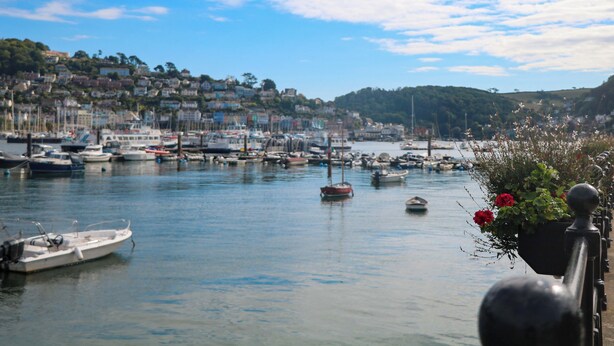 A harbor with boats, a waterfront promenade, and a hillside with buildings. - Full Deck (Dartmouth)