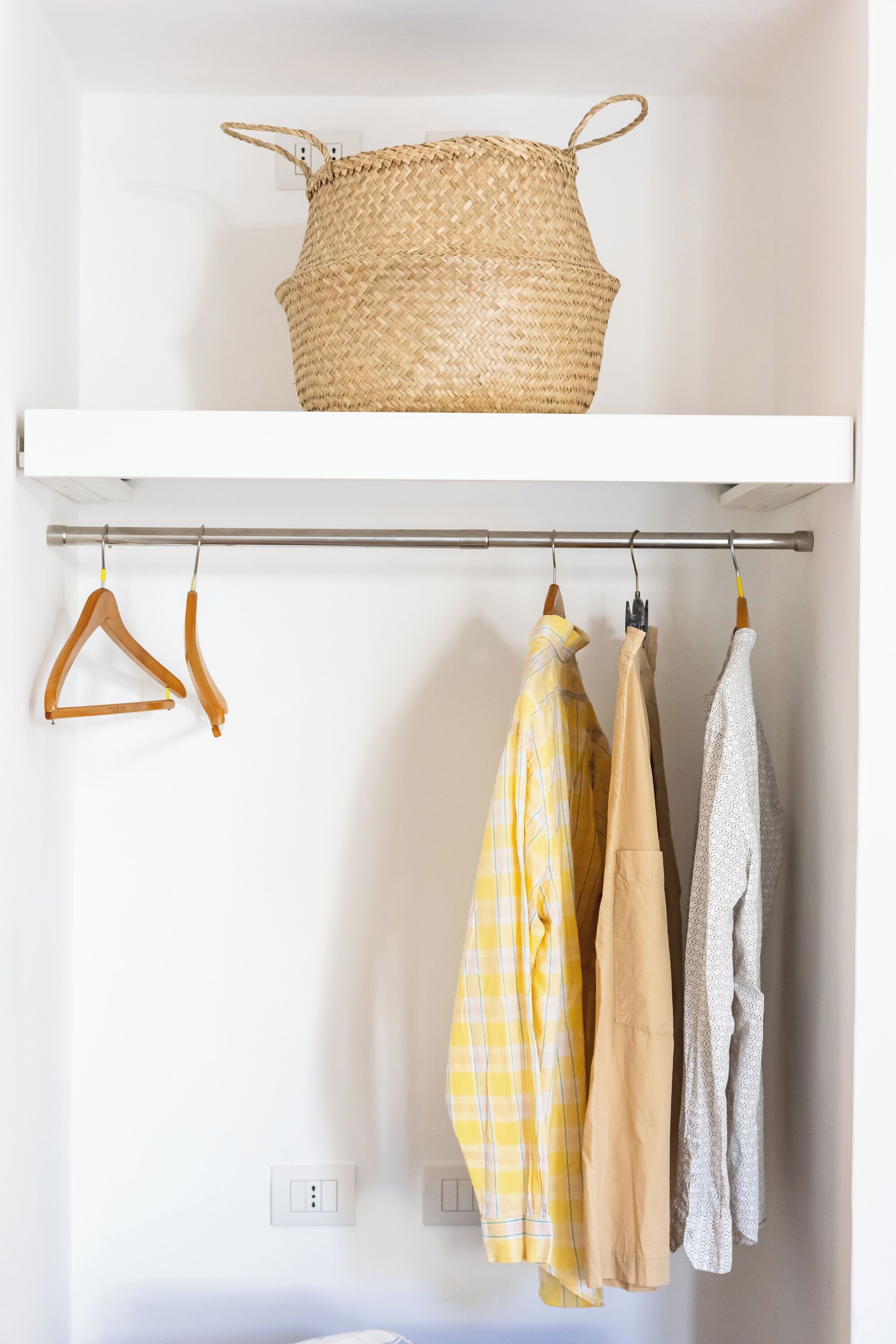A closet with a woven basket on a shelf, wooden hangers, and three shirts hanging on a rod.