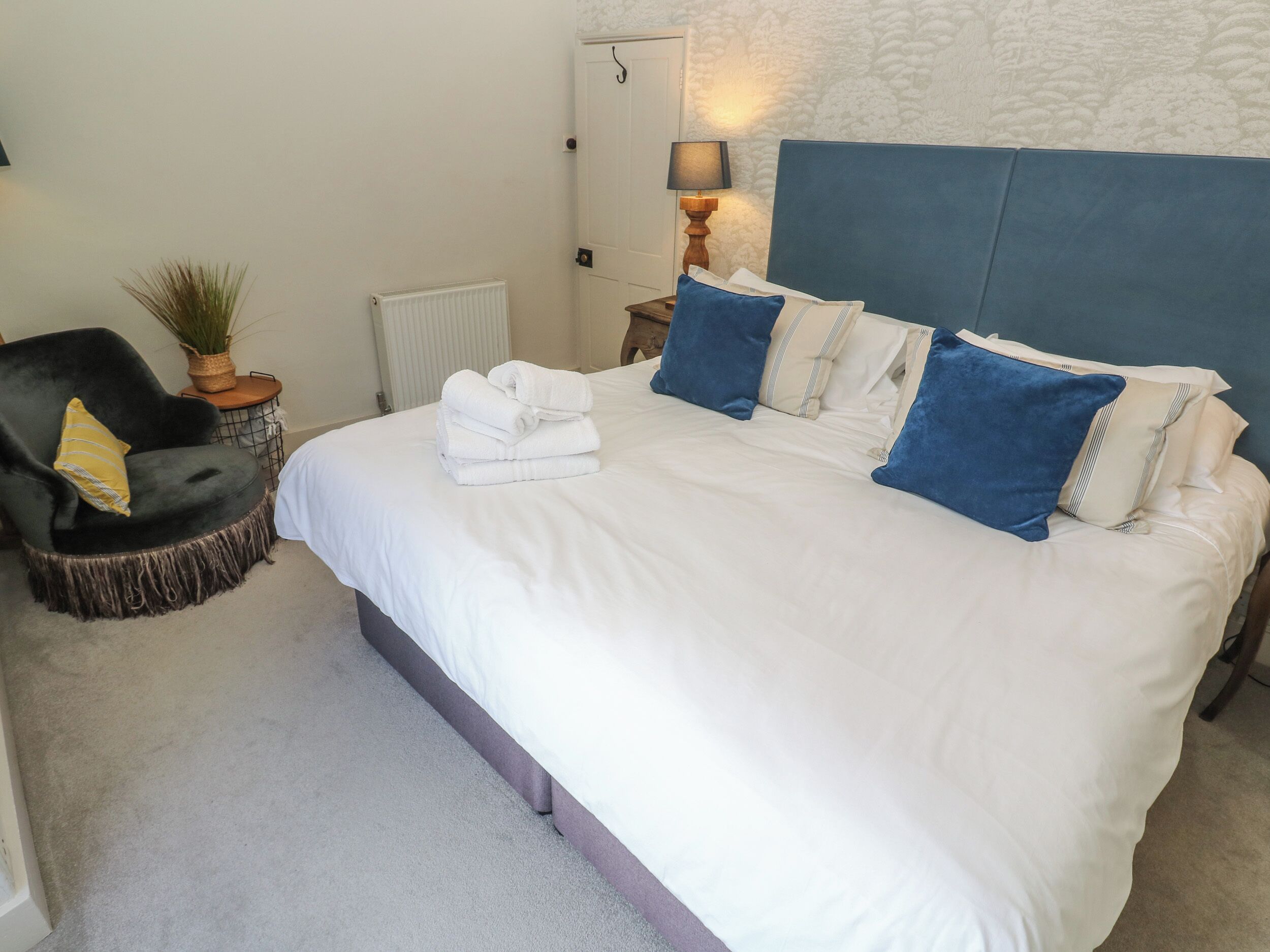 A neatly made bed with white linens and blue pillows, a dark armchair with a yellow cushion, a small side table with a lamp and a potted plant, and a radiator on the wall.