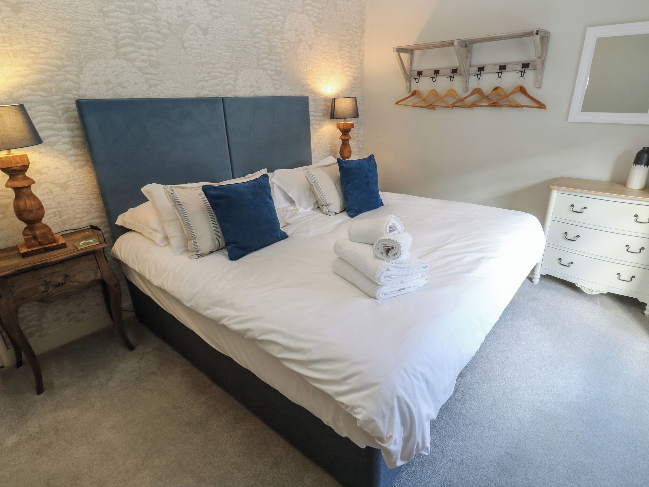 A neatly made bed with white linens and blue pillows, a bedside table with a lamp, and a dresser with drawers.