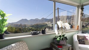 Ein Zimmer mit Blick auf Berge, ein Fensterbrett mit einem Topfpflanzen, ein kleiner Tisch mit einem roten Buch und ein weißer Stuhl.