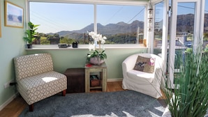 Ein gemütliches Zimmer mit großem Fenster und Blick auf die Berge, ein gemustertes Sessel, ein kleiner Tisch mit einer Pflanze und ein weißer Stuhl mit Kissen.