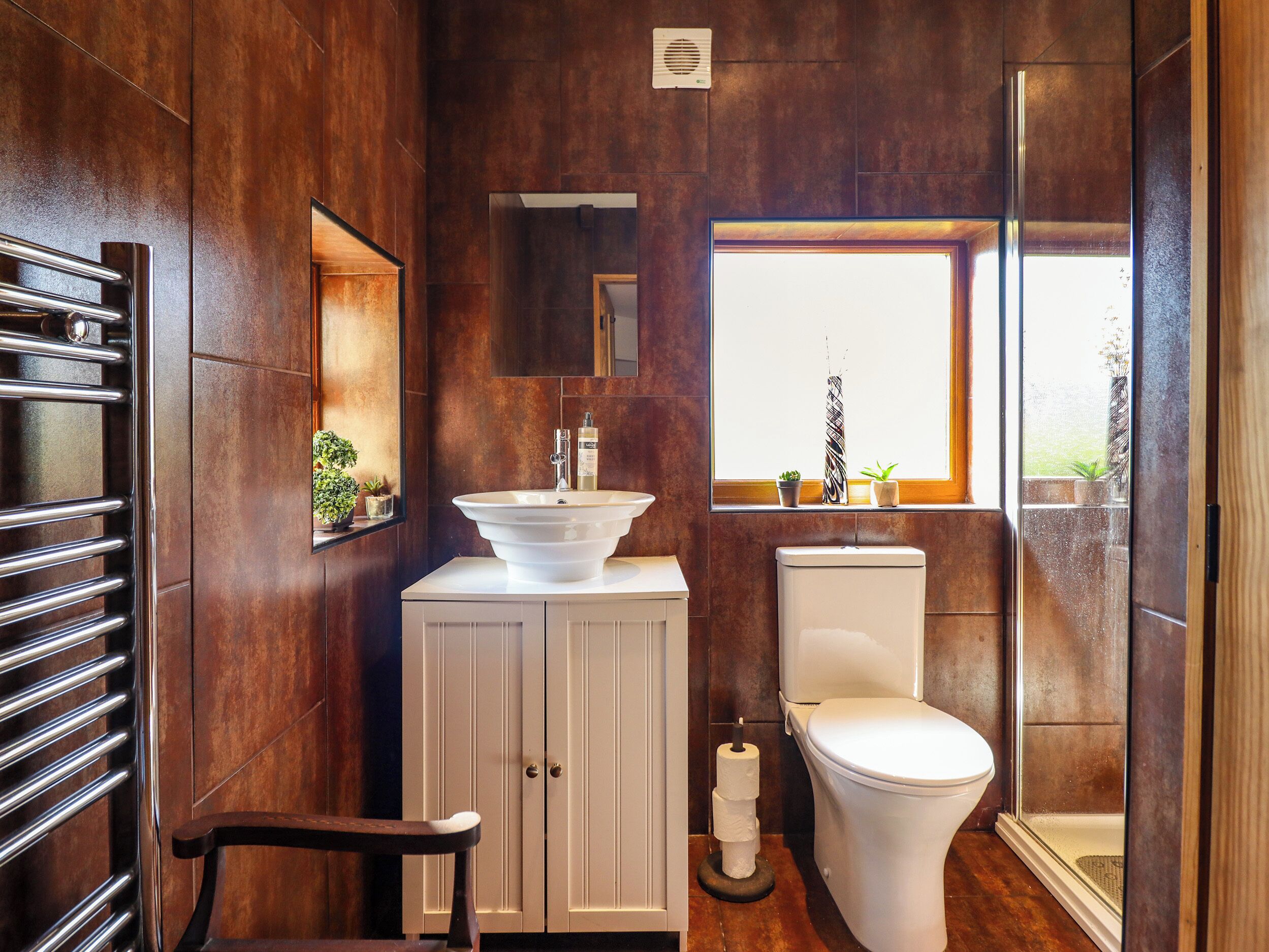 A bathroom with a white sink, a toilet, a shower, and a mirror. There are plants on the windowsill and a towel rack.