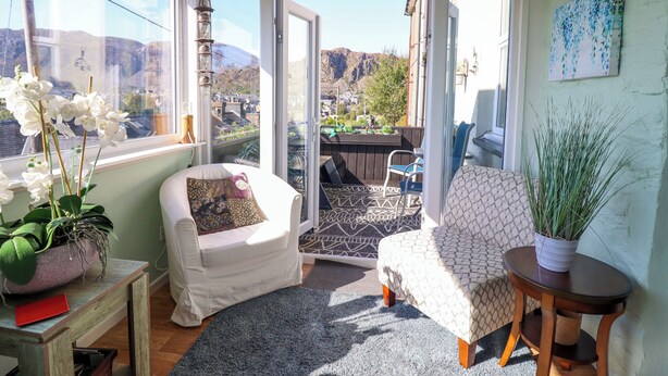 Ein gemütliches Zimmer mit Blick auf eine gebirgige Landschaft, ein weißer Sessel, ein kleiner Tisch mit einer Pflanze und ein gemustertes Teppich.