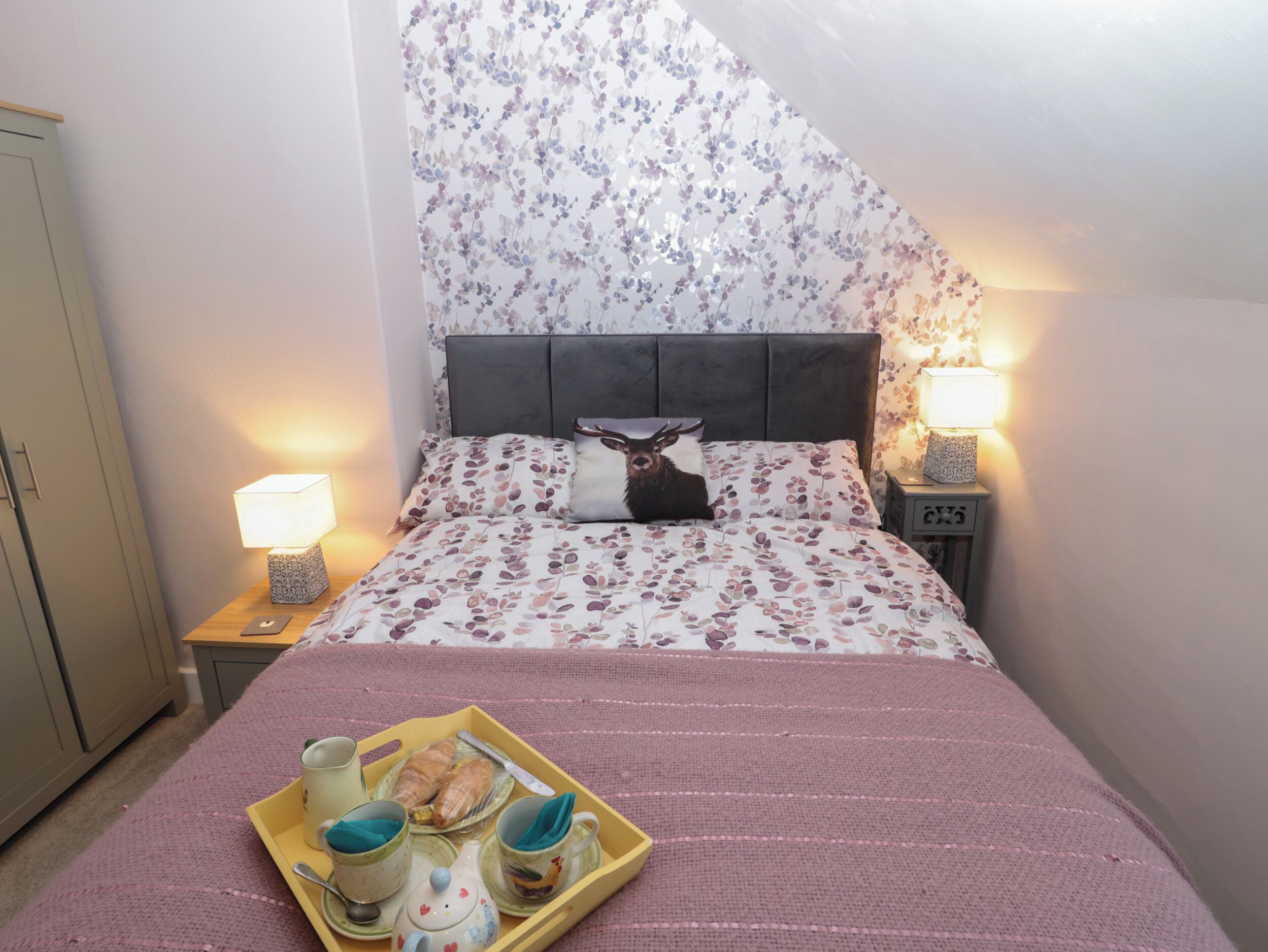 A bedroom with a bed, floral wallpaper, a headboard, bedside lamps, a nightstand, a wardrobe, and a tray with tea cups and pastries.