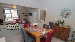 A dining area with a wooden table set for four, surrounded by red chairs. A sofa and a television are visible in the background. - Skyelands (Wooler)