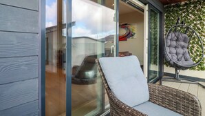 A wicker chair with a cushion on a balcony with glass doors and a hanging chair.