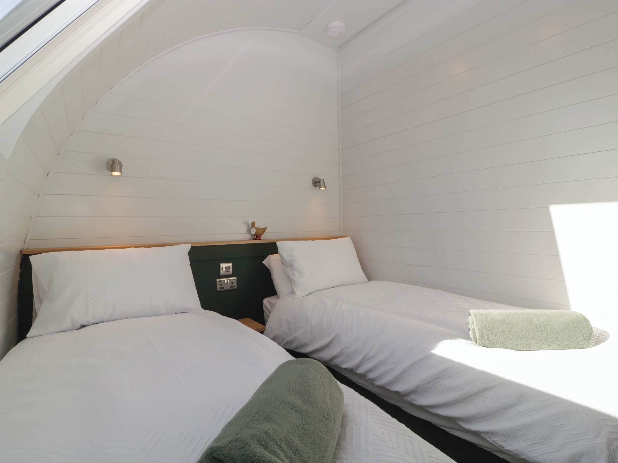 A bedroom with two beds, a skylight, a small table with a decorative item, and a wall-mounted light fixture.