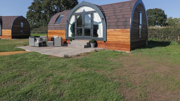 A modern, single-story house with a curved roof and large windows. It features a patio with outdoor furniture and a small garden area. - Leadon View Willow (Newent)