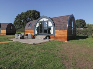A modern, single-story house with a curved roof and large windows. It features a patio with outdoor furniture and a small garden area. - Leadon View Willow (Newent)