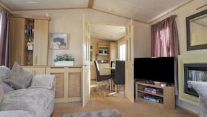 A cozy living room with a sofa, a TV, a bookshelf, and a kitchen area visible through an open door. - Ashcroft (Preston)