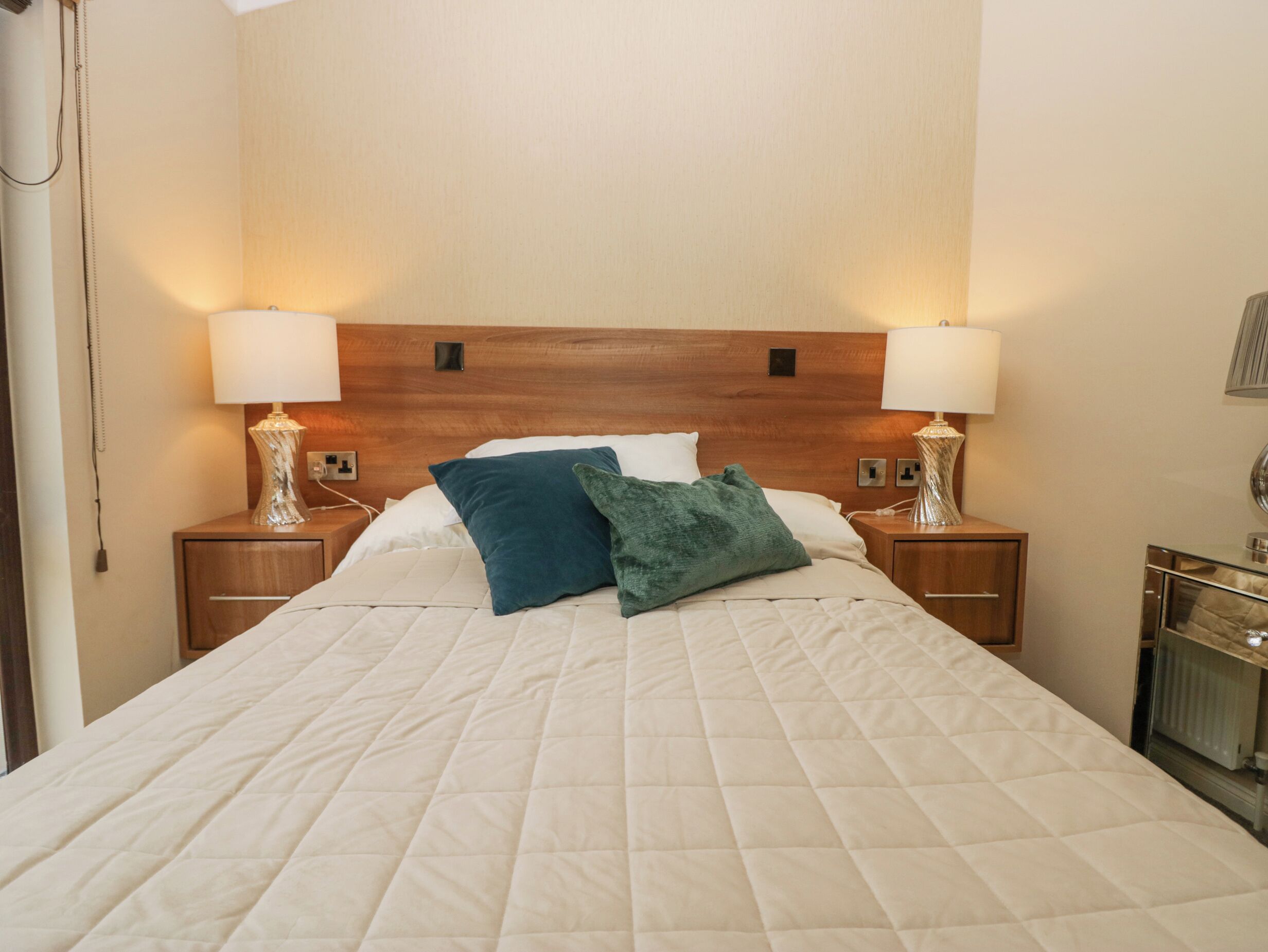 Cama bem arrumada com cobre acolchoado, duas luminárias de cabeceira e cabeceira de madeira.
