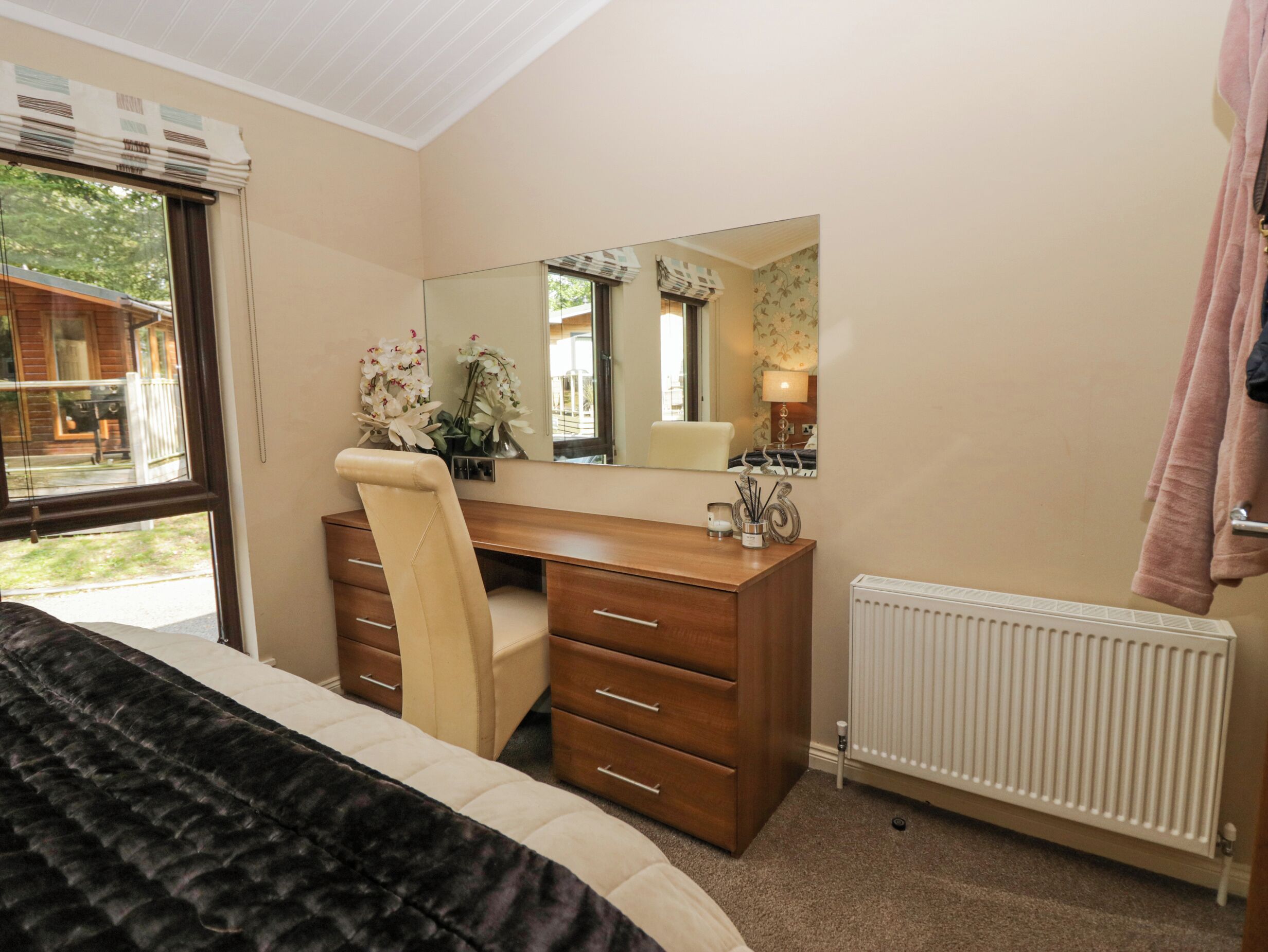 A bedroom with a bed, a wooden desk, a chair, a dresser, a radiator, and a window with a view of a house and trees.