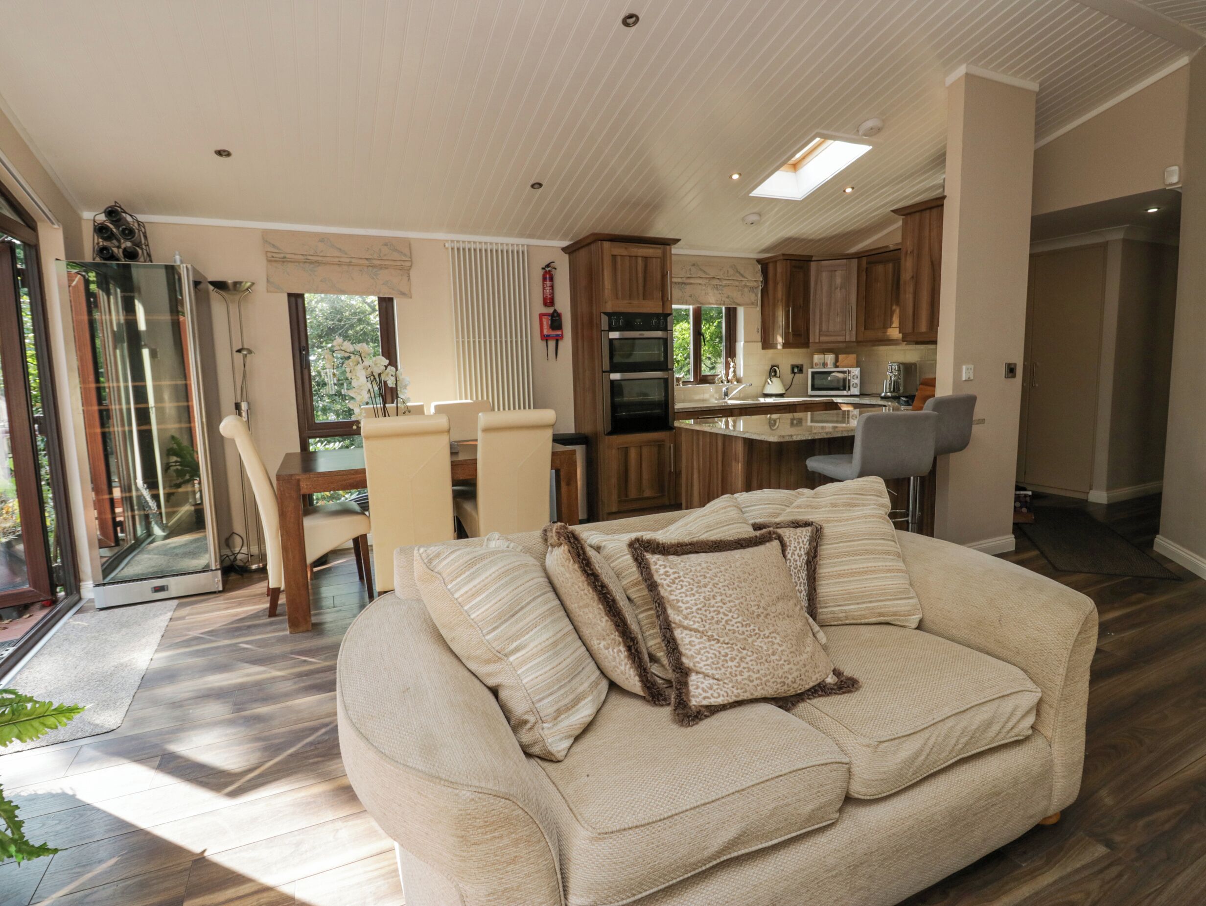 Een moderne woonkamer met een beige bank, een eethoek met een glazen deur naar een terras, en een keuken met houten kasten en een centraal kookeiland.