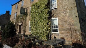 Un edificio de piedra con hiedra cubriendo parte de la fachada, bancos de madera y un cartel que dice "The Rose & Crown".