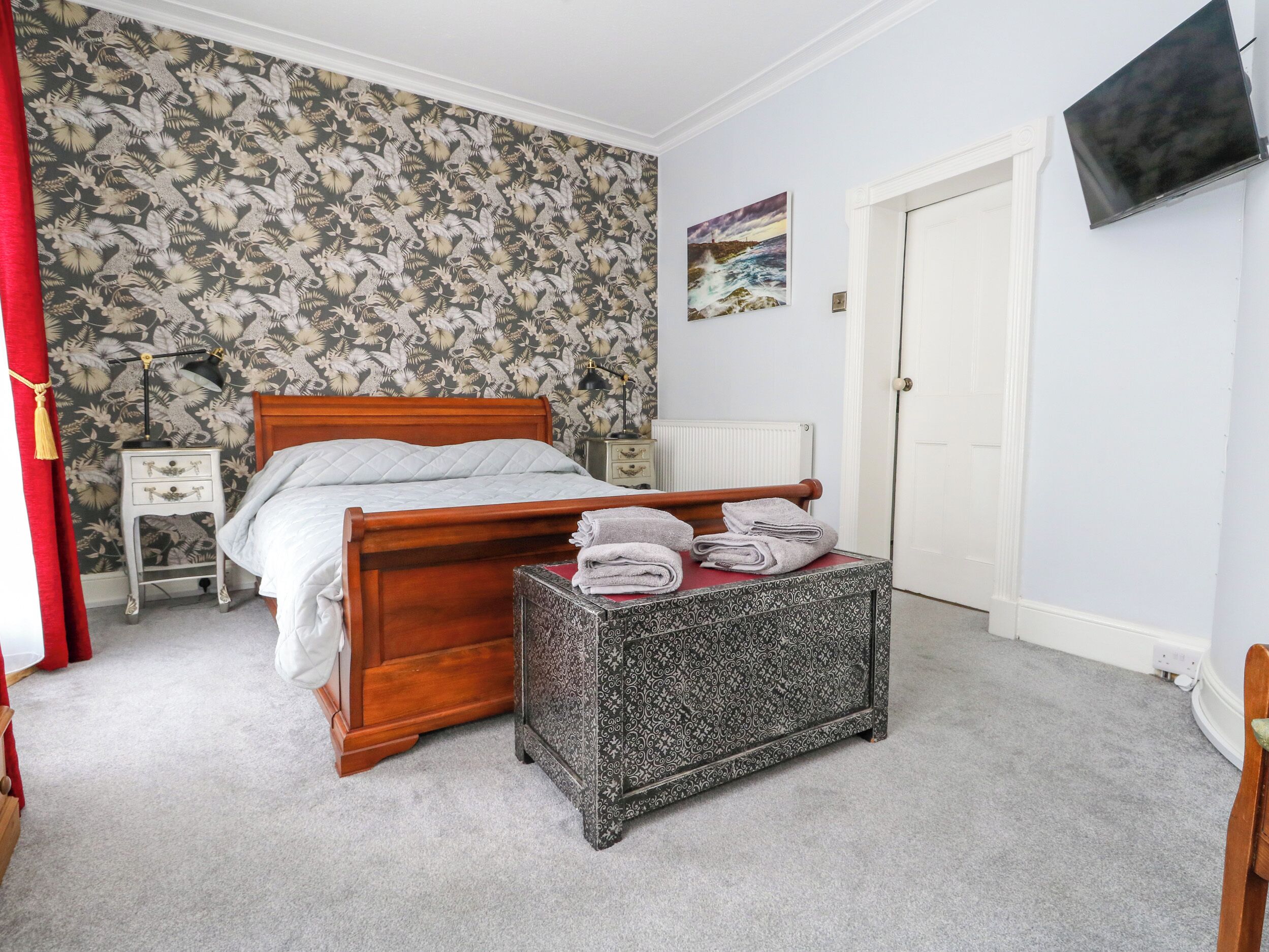 A bedroom with a wooden bed, a patterned ottoman, a nightstand, and a wall-mounted TV.