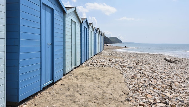 A row of blue beach huts along a pebbled beach with the sea in the background. - Charleston Cottage (Bridport)