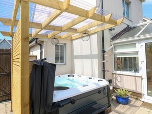 A hot tub under a wooden pergola with a clear roof, a potted plant, and a window with a plant inside. - Alban Road (Llanelli)