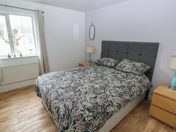 A bedroom with a bed, a nightstand, a lamp, a window with curtains, and a radiator. - Alban Road (Llanelli)