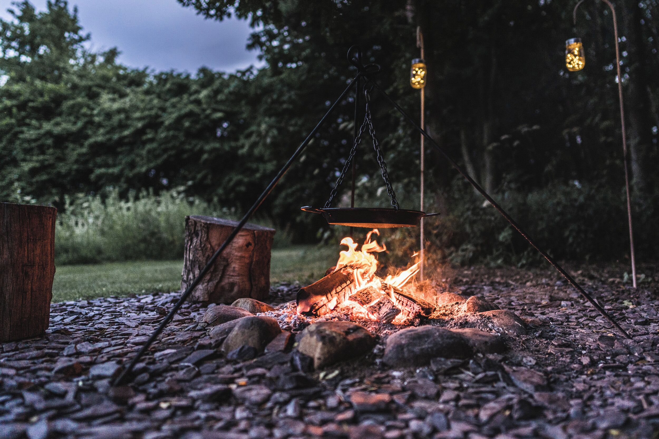 A campfire with logs and a metal bowl, surrounded by rocks and trees.