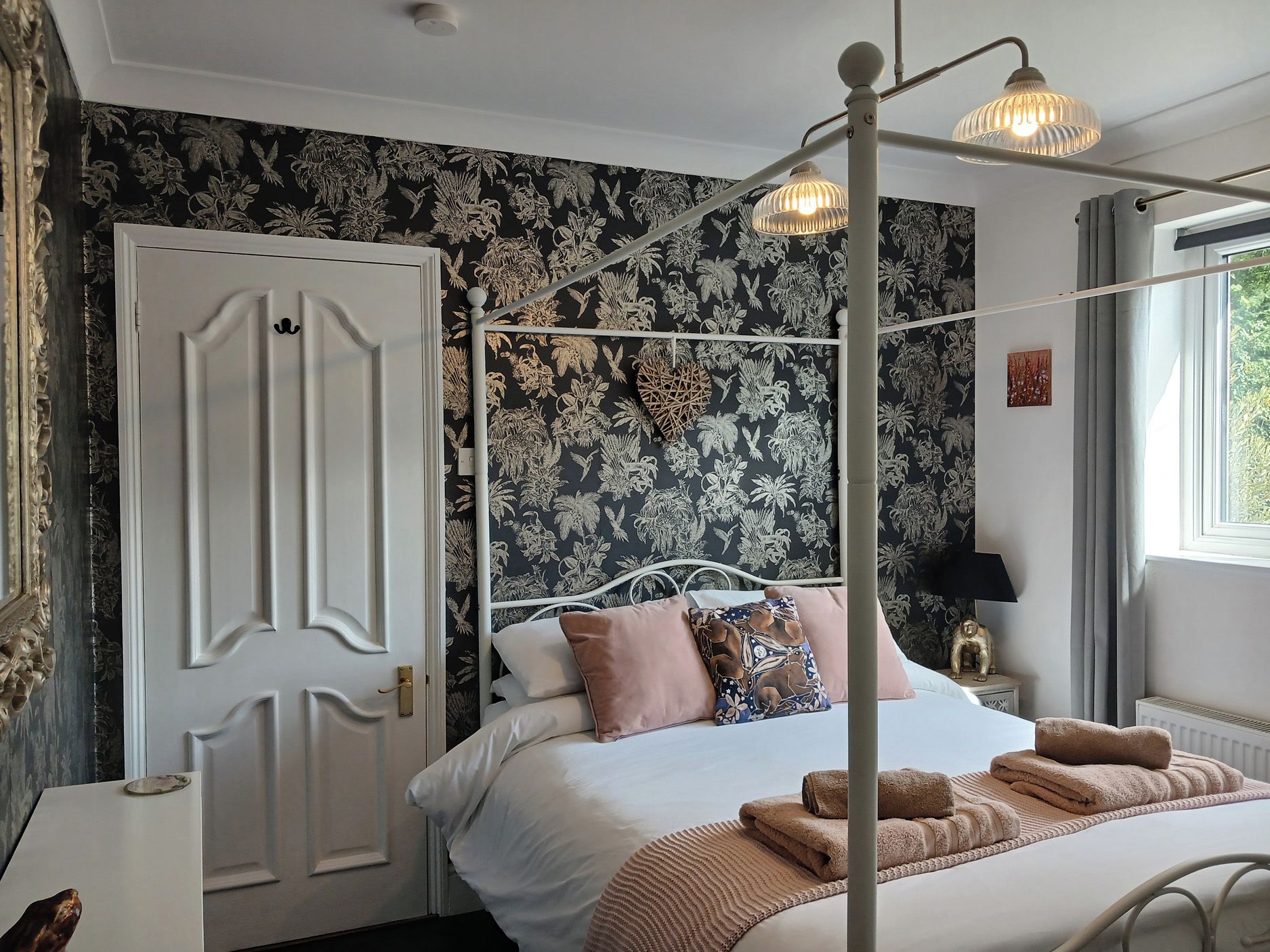 A bedroom with a four-poster bed, floral wallpaper, a white door, and a window with curtains.