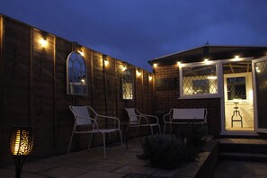 An outdoor patio area with wooden walls, two white chairs, a small table, and a decorative lamp. - The Corner House (Woodbridge)