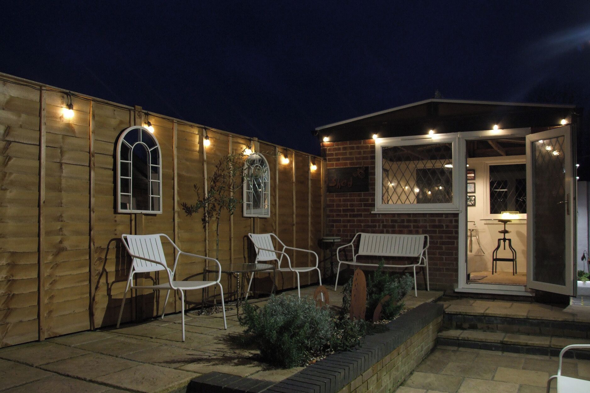 An outdoor patio area with white metal chairs and tables, a brick wall, and a glass door leading to an interior space.