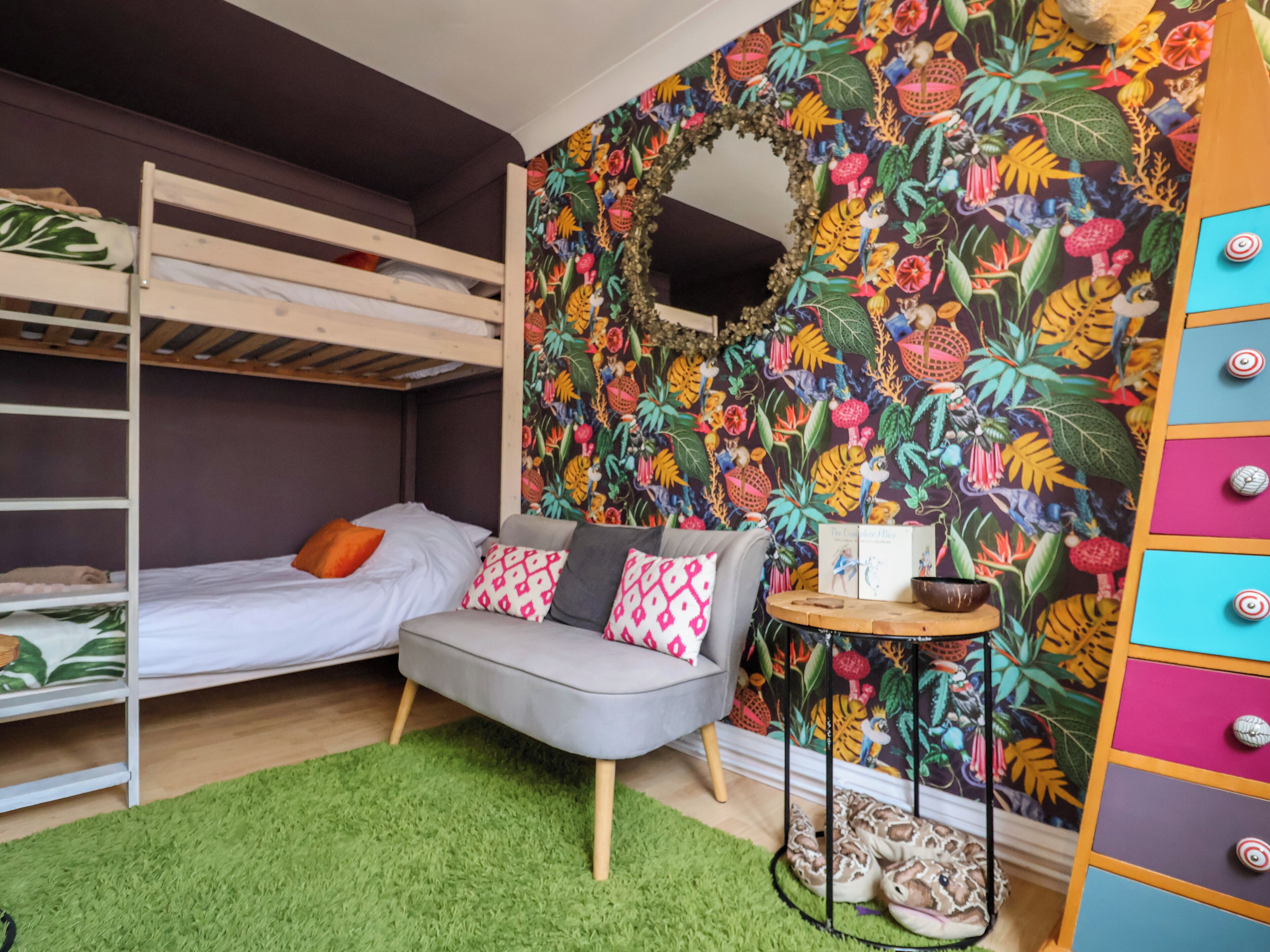 Una cama litera con un sofá gris, una mesita con libros y un papel tapiz tropical y colorido.
