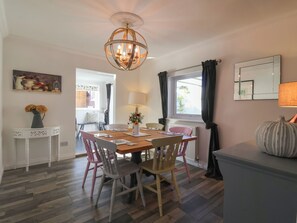 A dining area with a wooden table, chairs, a chandelier, a vase with flowers, a painting, and a window with curtains. - The Corner House (Woodbridge)