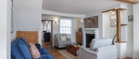 A living room with a blue sofa, a white armchair, and a fireplace. There is a wooden ceiling beam and a window with blinds.