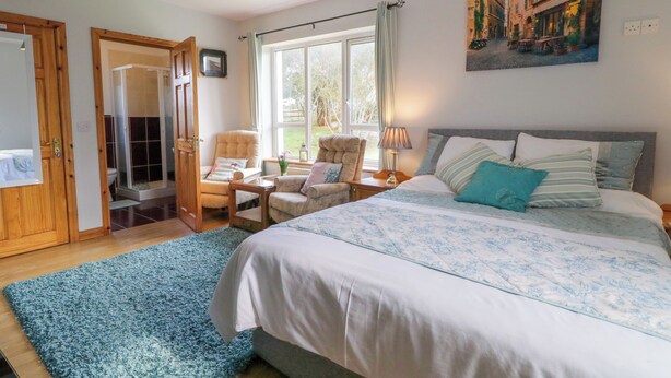 A bedroom with a large bed, two armchairs, a window with curtains, a painting on the wall, and a wooden door. - Thomond Hill (Naran)
