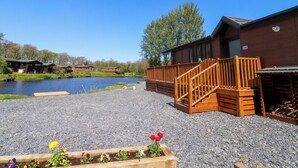 A wooden cabin with a gravel patio, a wooden deck, and a pond in the background.