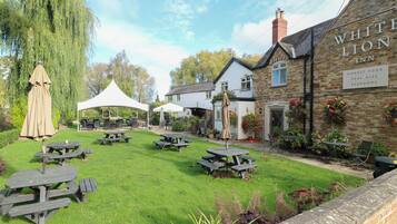 Un espace jardin avec des tables de pique-nique et une tente blanche, un bâtiment en pierre portant l’inscription « WHITE LION INN » et une pancarte indiquant « NOURRITURE FRAÎCHE, BIÈRES ARTISANALES, CHAMBRES ».