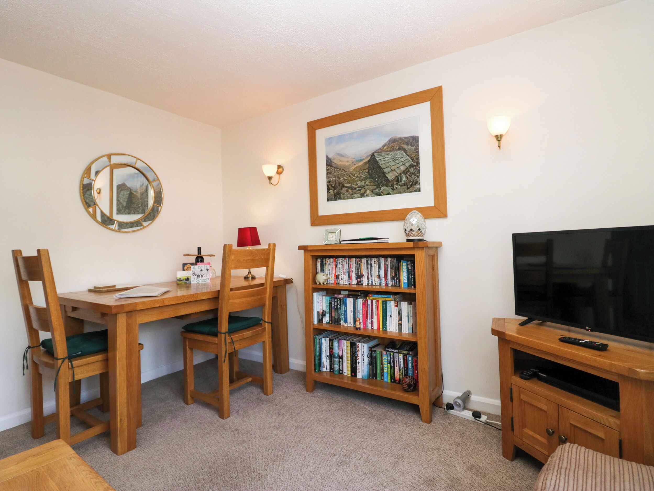 A cozy living room with a wooden dining table, two chairs, a bookshelf, a framed landscape painting, a television, and a lamp.