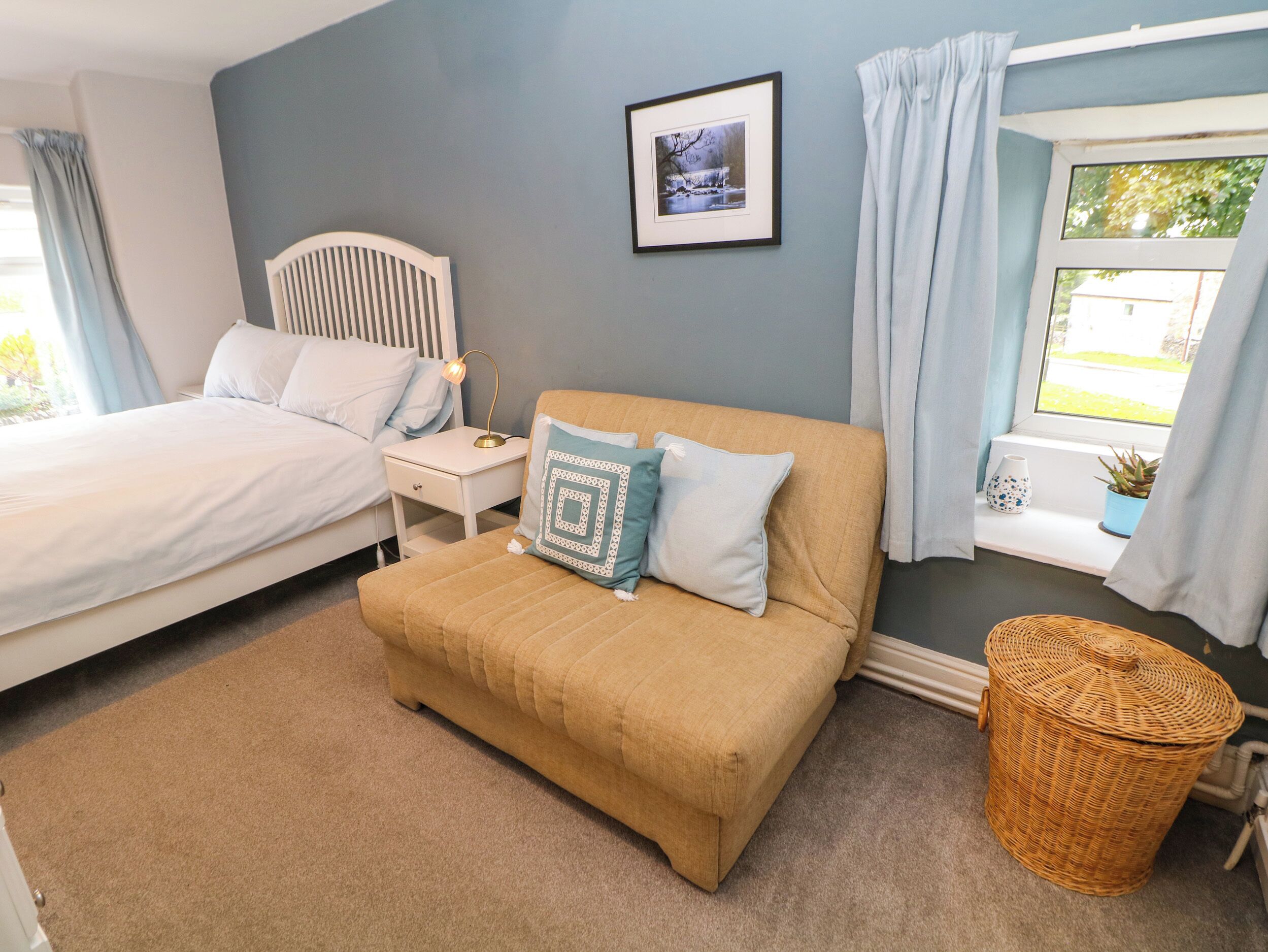 A bedroom with a bed, a sofa, a nightstand, a lamp, a framed picture, a window with curtains, and a wicker basket.