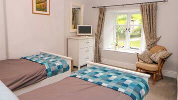 A bedroom with two single beds, a TV on a dresser, a chair with patterned cushions, and a window with curtains.