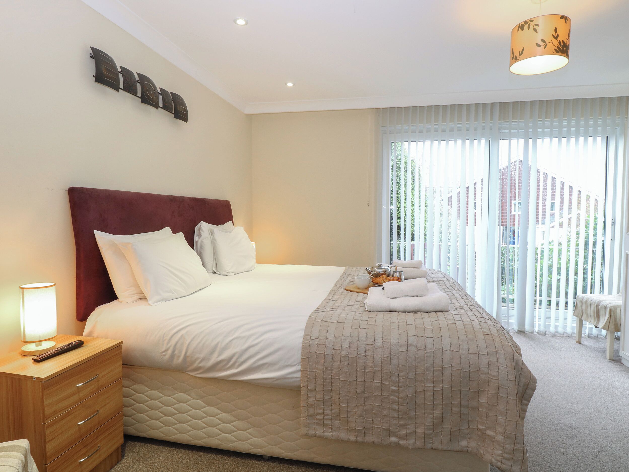 A neatly made bed with white linens, a wooden nightstand with a lamp, and a window with blinds.