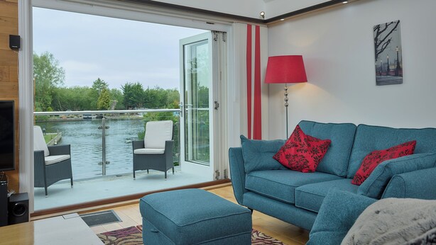 Ein modernes Wohnzimmer mit einem blauen Sofa, roten Kissen und einer Glastür, die auf einen Balkon mit Blick auf ein Gewässer führt.