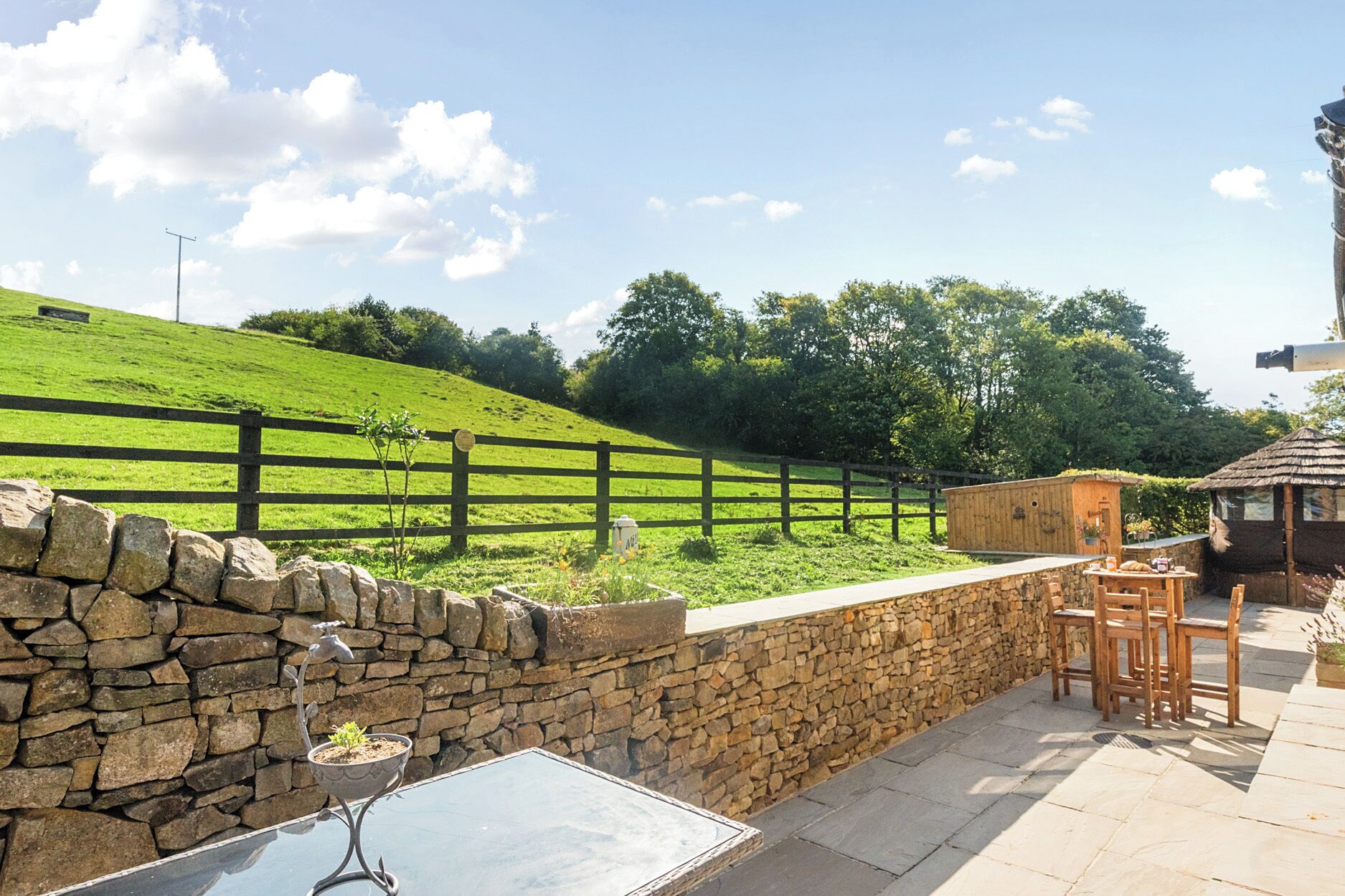 A stone wall with a table and chairs, a wooden fence, and a view of a green hill.