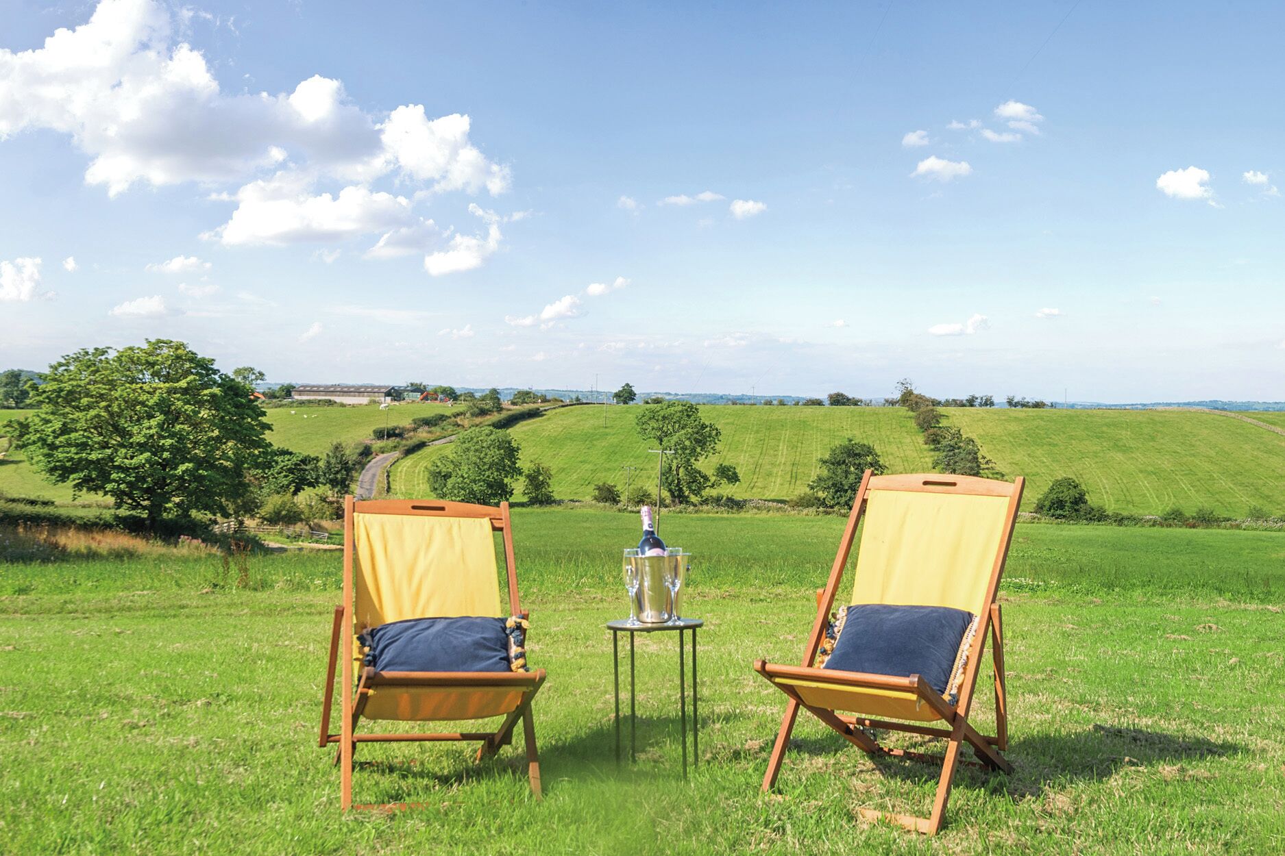Two wooden deck chairs with cushions, a bottle of champagne, and a small table on a grassy field with a view of rolling hills and a clear sky.