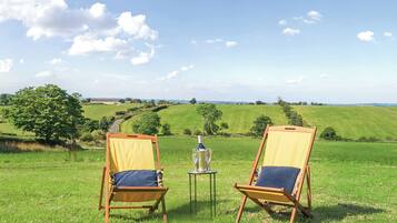 Two wooden deck chairs with cushions, a bottle of champagne, and a small table on a grassy field with a view of rolling hills and a clear sky.